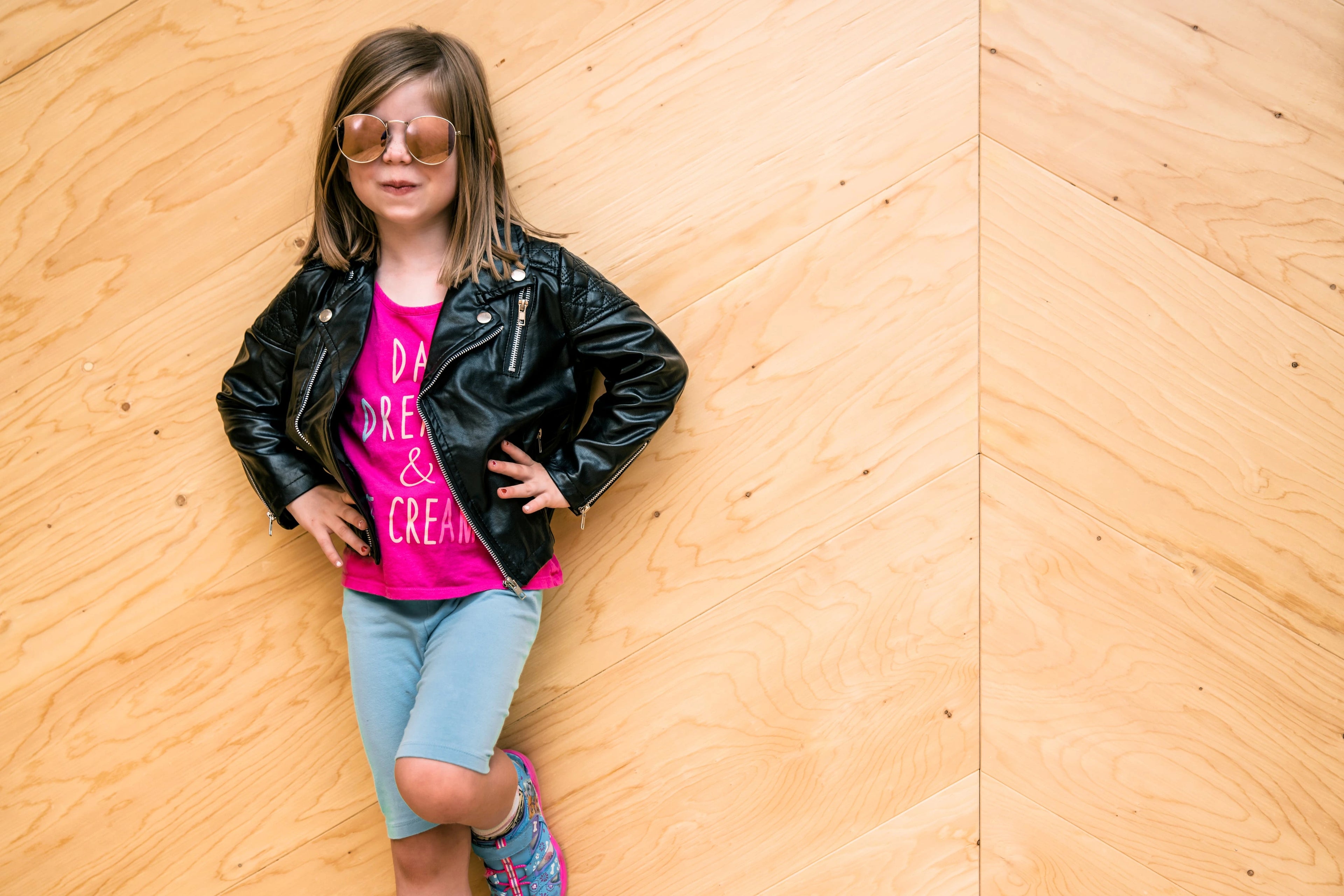 Young girl wearing a black leather jacket, pink shirt, and blue shorts against a wooden wall.